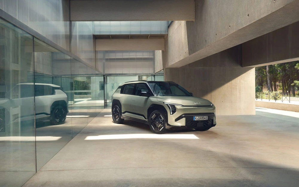 Açık gri renkteki modern ve köşeli hatlara sahip Kia EV3 SUV elektrikli otomobil, şık cam ve betonarme bir galerinin içinde park etmiş halde, dış mekan tasarımı detaylarıyla birlikte görülüyor; soldaki cam duvarda ise aracın yansıması belirginleşiyor.