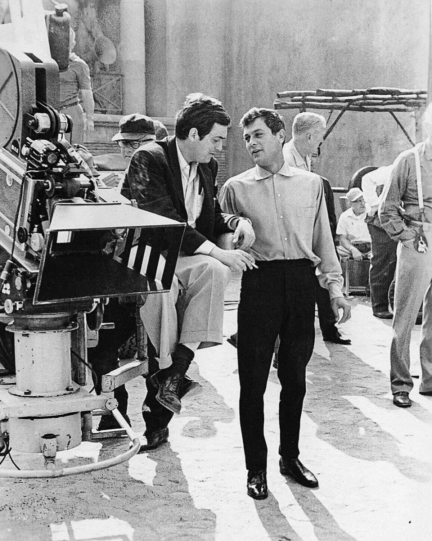 Yönetmen Stanley Kubrick, 1960 yapımı Spartacus filminin setinde büyük bir sinema kamerasına oturmuş veya yaslanmış vaziyette, oyuncu Tony Curtis ile güneşli bir dış mekanda sohbet ederken görülüyor.