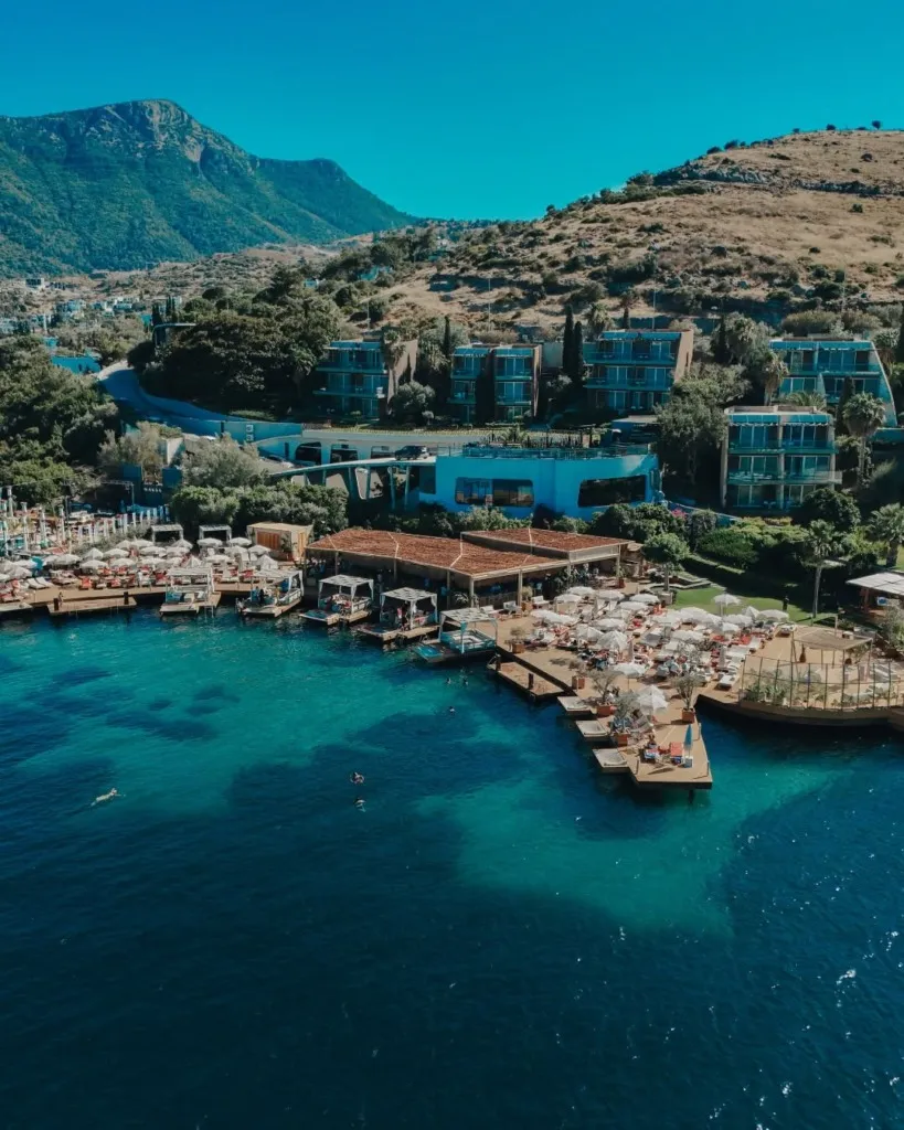Bodrum'da, Kuum Beach'in masmavi denizine uzanan iskeleleri ve şemsiyelerle dolu plaj alanları, arkasındaki modern otel binaları ve yeşil tepelerle bir arada, yukarıdan çekilmiş nefes kesici bir görüntü.