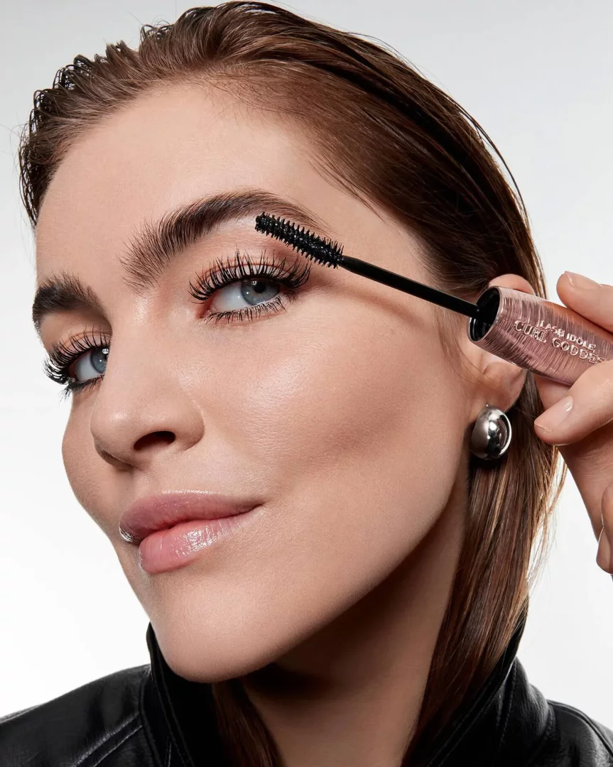 Mavi gözlü, ıslak görünümlü kahverengi saçlı ve siyah deri ceketli bir kadın, elindeki Lancôme Lash Idôle Curl Goddess maskara fırçasıyla belirgin, uzun ve kıvrık kirpiklerine son rötuşları yaparken, ürünün kirpiklere anında hacim ve kalkıklık kazandırdığı açıkça görülüyor.
