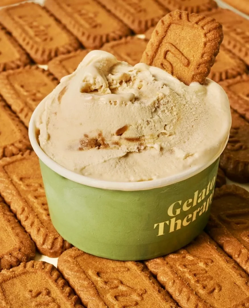 Altın sarısı Gelato Therapy yazılı yeşil bir kapta, içine diklemesine batırılmış bir Lotus Biscoff kurabiyesiyle süslenmiş, etrafı düzgün sıralanmış çok sayıda Lotus Biscoff kurabiyesiyle çevrili, kremalı ve parçacıklı Lotus Biscoff gelato iştah açıcı bir şekilde sergileniyor.