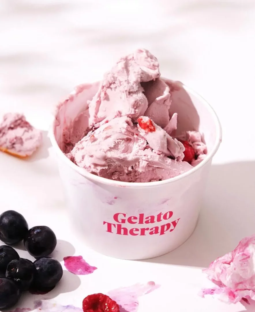 Beyaz bir masa üzerinde, pembe tonlarında, taze ahududu ve yaban mersini parçacıklarıyla zenginleştirilmiş, üzerinde pembe harflerle Gelato Therapy yazan beyaz bir kapta servis edilen bol kepçeli dondurma, etrafına dağılmış canlı renkli yaban mersinleri, bir frambuaz ve arka planda küçük bir külah parçası üzerindeki erimiş dondurma kalıntısı ile iştah açıcı bir yaz lezzetini gözler önüne seriyor.