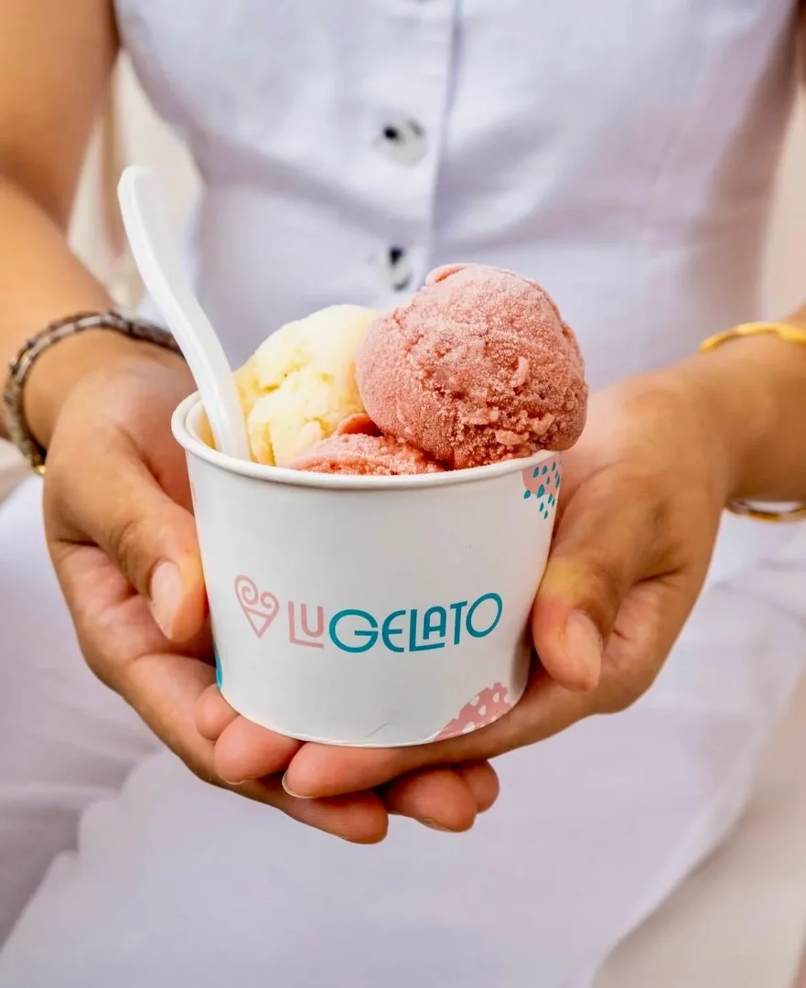 Beyaz kıyafetli bir kişinin ellerinde, üzerinde Lu GELATO logosu bulunan beyaz bir kap içinde, biri sarı diğeri pembe renkli iki top gelato ve beyaz bir kaşık görülüyor; bu serinletici lezzet, sıcak havalarda ferahlatıcı bir mola sunuyor.