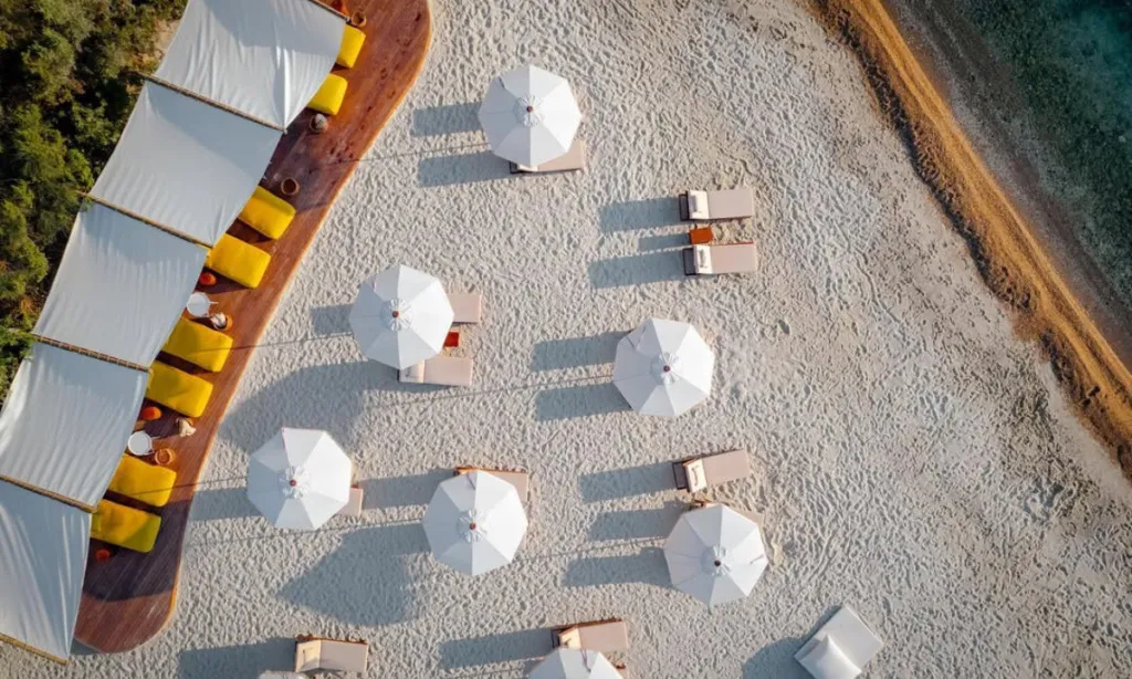Lucca Beach Bodrum Gölköy’de beyaz kum plaj, sarı minderli gölgelikler, şezlonglar ve turkuaz deniz manzarası