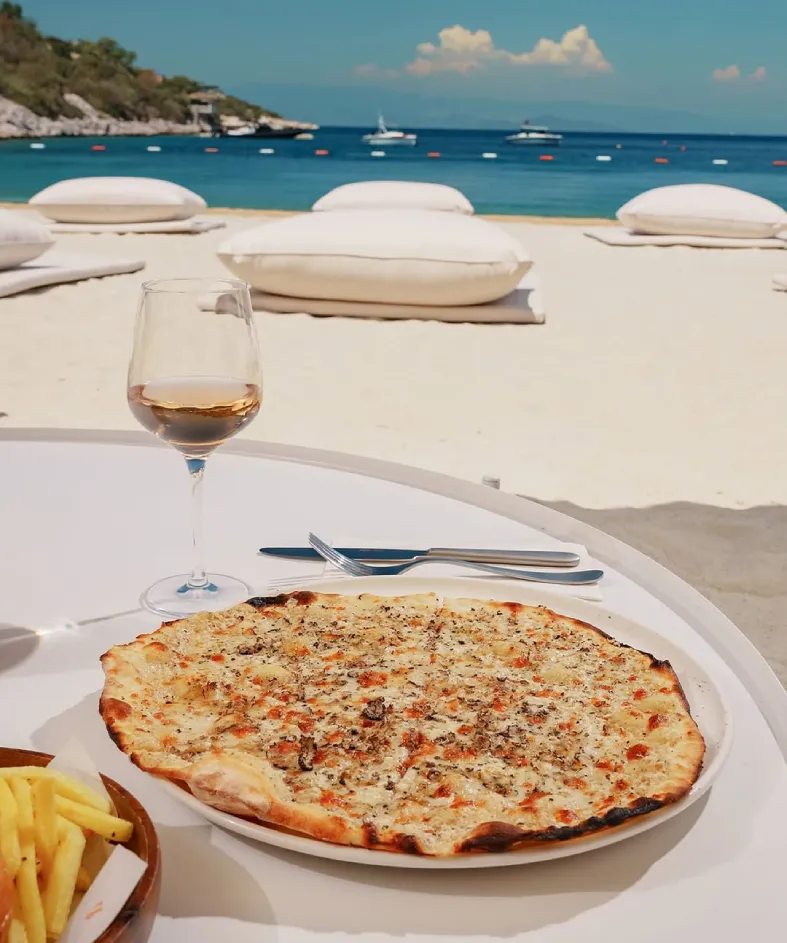 Beyaz kumlu plajda, üzerinde gurme bir ince hamur pide veya pizza, bir kadeh roze şarap ve patates kızartması olan beyaz bir masa, arkasında beyaz minderlerin uzandığı kumsalda teknelerle dolu masmavi deniz ve uzak yeşil sahil, Bodrum'da huzurlu bir sahil restoranı atmosferini gözler önüne seriyor.