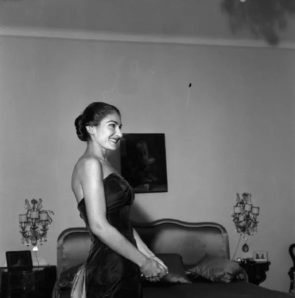 Maria Callas'ın 1957 Milano'da çekilmiş bu samimi siyah beyaz portresinde, zarif topuzlu saçları ve inci gibi dişleriyle parlayan bir gülümsemeyle straplez, koyu renk bir gece elbisesi içinde, elleri kenetli bir şekilde lüks bir yatak odasında duruşu dikkat çekiyor.