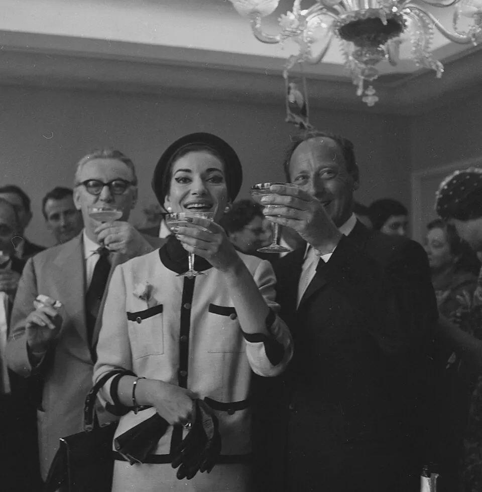 Siyah beyaz bir fotoğrafta, koyu renk beresi ve açık renk ceketiyle göz alıcı soprano Maria Callas, bir kadeh şampanyayla neşeyle gülümserken yanında koyu takım elbiseli bir adam da kadehini kaldırmış, arka planda diğer konuklar ve bir avizeyle dolu bu kutlama anı, ünlü opera sanatçısının başarılı bir etkinliğinin ardından çekilmiş gibi görünüyor.