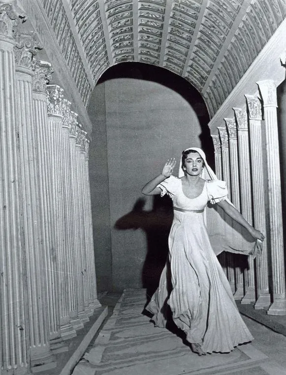 Maria Callas, açık renk pilili uzun elbisesi ve başını örten hafif bir örtüyle, antik temalı sütunlu bir koridor ile süslü kemerli bir tavanın altında, elini havaya kaldırmış, endişeli bir yüz ifadesiyle dramatik bir sahne performansı sergiliyor.