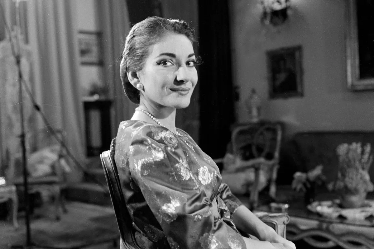 20. yüzyılın zamansız opera ikonu Maria Callas, inci küpeleri ve kolyesiyle, desenli kıyafeti içinde zarif bir şekilde otururken, arkasına dönerek kameraya hafifçe gülümseyen, siyah beyaz bir portre fotoğrafı.