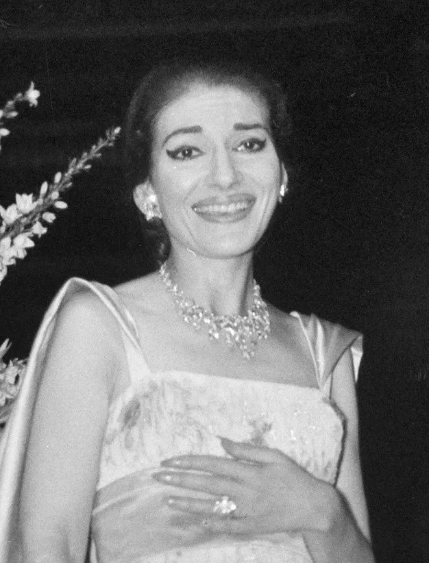 Maria Callas'ın 1959 Amsterdam'da bir etkinlikte çekilen bu siyah beyaz portresinde, sahne makyajı, pırıltılı pırlanta kolyesi, küpeleri ve yüzüğüyle dikkat çeken zarif bir elbise içinde, geniş bir gülümsemeyle kameraya bakarak coşkuyla poz verdiği görülüyor.