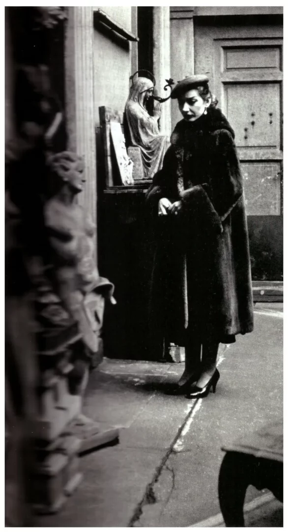 Maria Callas, 1955 yılında Milano'da çekilen bu siyah beyaz fotoğrafta, koyu renk kürk mantosu ve şık şapkasıyla çatlak zemin üzerinde, arka planda dua eden bir figür ve ön planda bulanık bir heykelin önünde düşünceli bir ifadeyle ayakta duruyor.