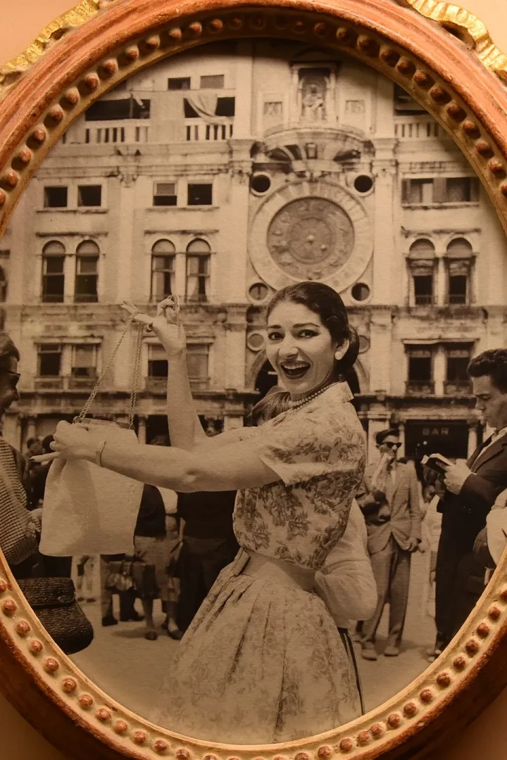Maria Callas'ın Venedik'te, ünlü San Marco Saat Kulesi'nin önünde, inci kolyesi ve desenli elbisesiyle neşeli bir şekilde gülümserken el çantasını tuttuğu, sahne dışı bir anını yakalayan siyah beyaz fotoğraf.