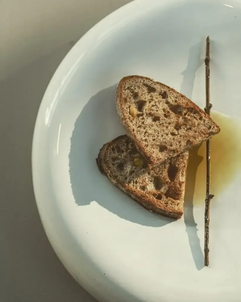 Beyaz ve minimalist bir tabağın üzerinde, güneşin yumuşak ışıkları altında duran iki dilim rustik tam tahıllı ekmek, sağ tarafında ince bir ağaç dalı ve ekmeğin altında hafifçe yayılmış zeytinyağına benzeyen sarımsı bir sıvı ile mezra yaşamının doğallığını ve sakinliğini vurgulayan sade bir görsel.