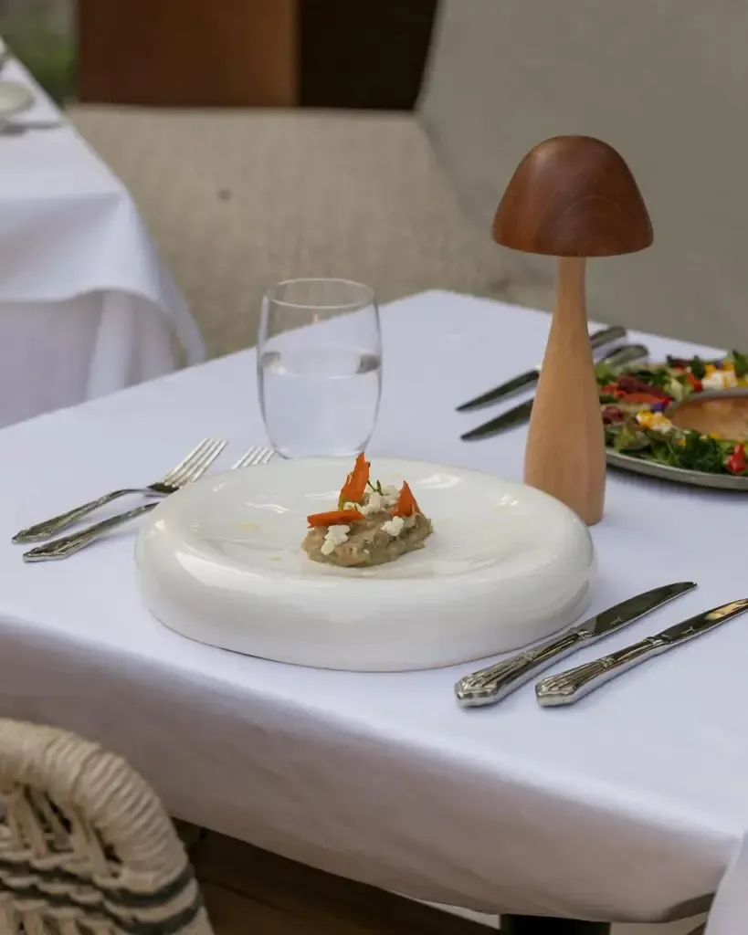 Beyaz masa örtülü şık bir restoranda, yuvarlak hatlı beyaz bir tabakta havuç dilimleri, beyaz peynir parçacıkları ve taze otlarla süslenmiş bir meze veya başlangıç yemeği, yanında zarif çatallar, bıçaklar, su bardağı ve masaya sıcak bir atmosfer katan mantar şeklinde ahşap bir dekoratif lamba ile Mezra deneyimini vurgulayan bir görsel.