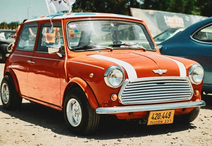 Kırmızı renkli klasik Mini Cooper Mk1, beyaz şerit detaylarıyla açık alanda sergileniyor. Kompakt tasarımıyla şehir otomobili kimliğini ve ralli mirasını yansıtıyor.