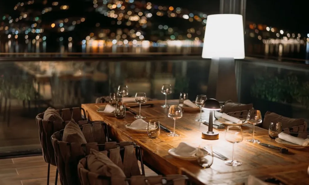 Gece, Bodrum'daki Mivara lüks otelin teras restoranında, ışıl ışıl şehir manzarasına bakan, zarif kadehler ve modern masa lambalarıyla donatılmış uzun ahşap bir yemek masası, sıcak ve davetkar bir ambiyans sunuyor.