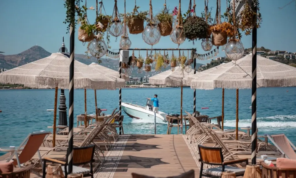 Bodrum seyahat rehberi: Bodrum'daki bir plajda ahşap bir iskelenin üzerinde, tığ işi detaylı beyaz şemsiyelerin gölgelediği şezlonglar ve bitki saksılarıyla cam kürelerin sarktığı dekoratif tavan süslemeleri bulunurken, masmavi denizde bir teknenin ilerlediği ve uzakta tepelerin yükseldiği panoramik bir yaz manzarası.