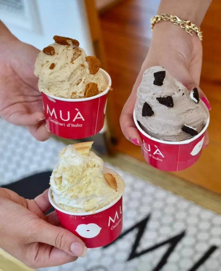 Birden fazla kişinin ellerinde, Biscoff kurabiyeli, Oreo kurabiyeli ve bisküvi kırıntılı olmak üzere üç ayrı lezzetteki MÙA İtalyan gelatoları kırmızı kaplarda tutuluyor; arka planda ahşap zemin ve markanın logosunu taşıyan desenli bir halı hafifçe görülüyor.