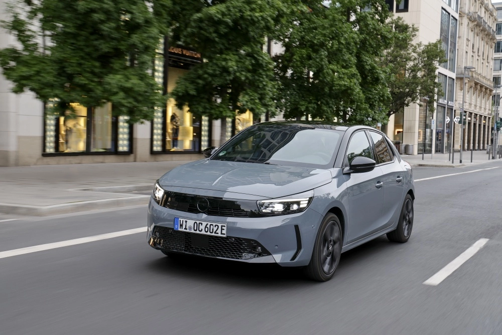 Gri rengi ve siyah tavan detaylarıyla dikkat çeken modern bir Opel Corsa Electric B-hatchback, parlak farları açık bir şekilde dinamik biçimde şehir caddesinde ilerlerken, markanın yeni elektrikli otomobil dış tasarım dilini sergiliyor.