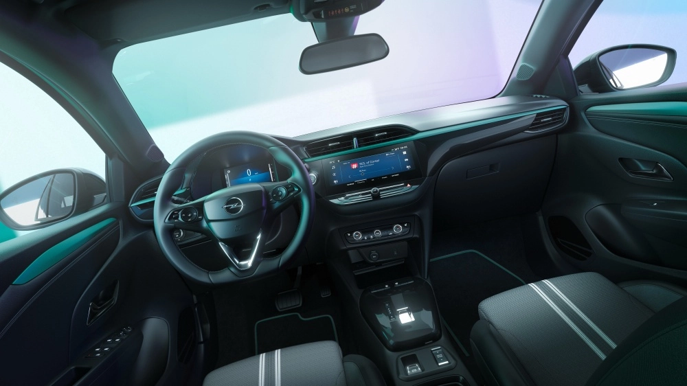 Opel Corsa Electric modelinin sürücü koltuğundan bakılan modern iç mekan tasarımı; dijital gösterge paneli, büyük multimedya ekranı, zarif orta konsolu, çizgili koltukları ve yeşil-mavi tonlarda ambiyans aydınlatmasıyla şık ve teknolojik bir elektrikli otomobil atmosferi sunuyor.