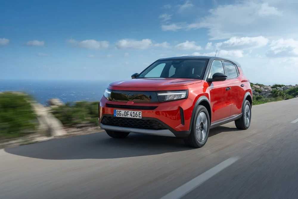 Kırmızı bir Opel Frontera Electric, masmavi denizin solunda ve kayalık yeşil tepelerin sağında uzanan manzaralı bir sahil yolunda ilerlerken, modern dış mekan tasarımı ve elektrikli yapısıyla dikkat çekiyor.
