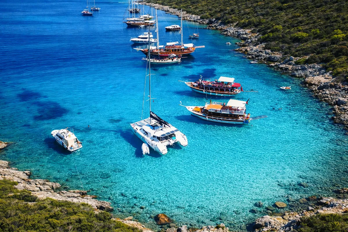 Bodrum seyahat rehberi: Bodrum'un gözde duraklarından Orak Adası'nın suları berrak turkuaz rengi olan muhteşem koyunda, farklı tiplerde birçok yelkenli, katamaran ve tur teknesi demirlemişken, bazı insanlar masmavi denizde yüzerek bu doğal güzelliğin tadını çıkarıyor ve kıyılar yemyeşil bitki örtüsüyle çevrili kayalıklarla yükseliyor.