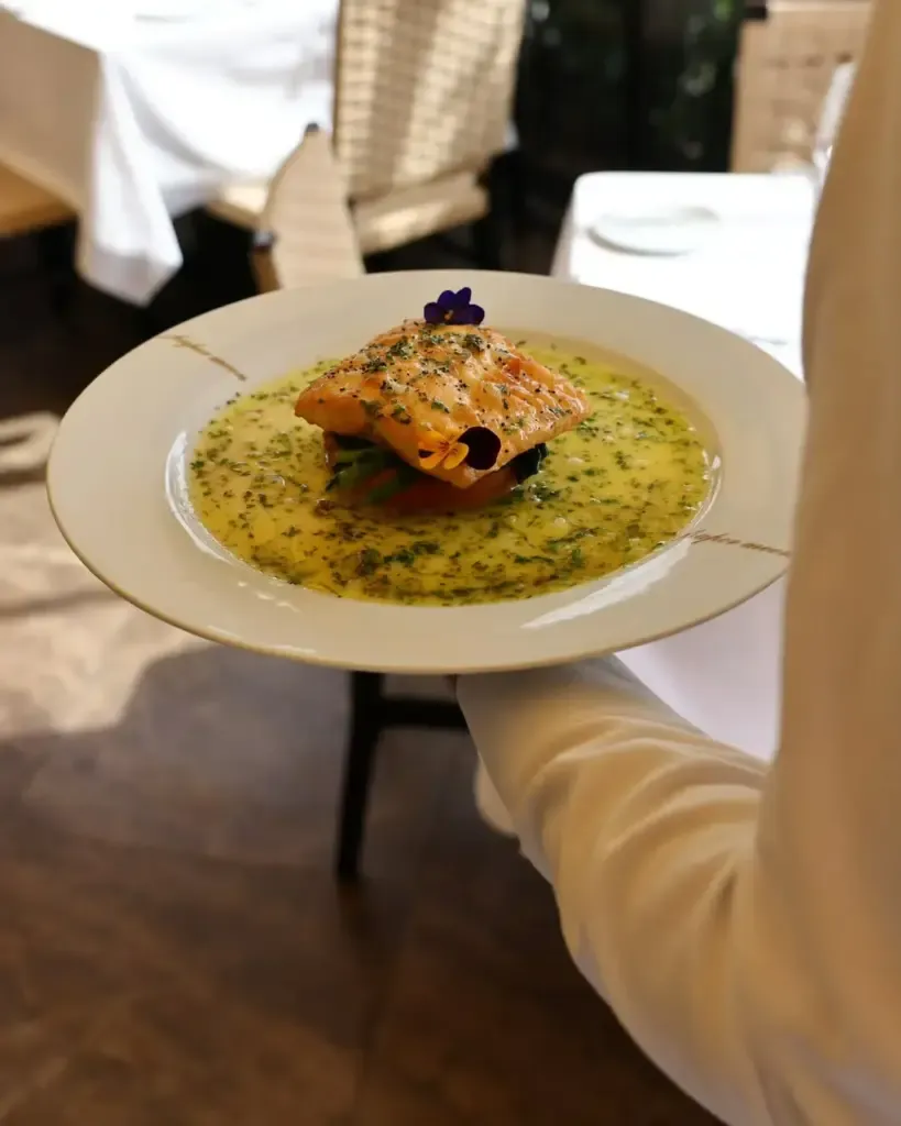 Beyaz üniformalı bir garsonun tuttuğu tabakta, Paper Moon restoranının özenle hazırlanmış somon yemeği sunuluyor; sarımsı otlu sos içinde sebzelerin üzerinde servis edilen kızarmış somon fileto, mor ve sarı yenilebilir çiçeklerle süslenmiş.