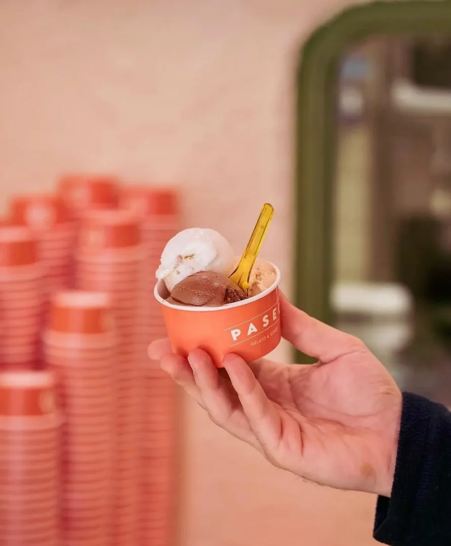 Bir el, içinde beyaz, çikolatalı ve açık kahverengi üç top dondurma ile sarı bir kaşık bulunan turuncu Paseo Gelato & Sorbet kupasını tutuyor; arka planda benzer turuncu kaplardan oluşan yığınlar ve yeşil bir ayna çerçevesi belli oluyor.