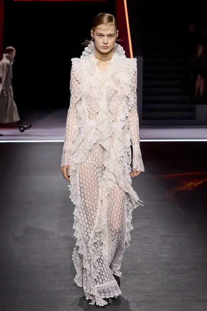 Katmanlı beyaz dantel uzun elbise içinde yürüyen model, romantik dokular ve heykelsi siluetlerle haute couture zarafetini öne çıkarıyor.