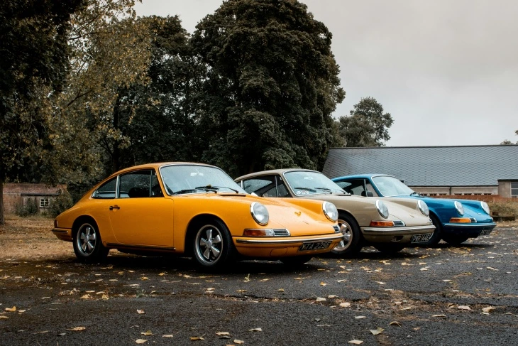 Sarı, bej ve mavi renklerde üç klasik Porsche 911 modeli, ağaçlarla çevrili bir alanda yan yana sergileniyor. İkonik tasarımıyla tanınan 911, otomobil tarihinin en zamansız modellerinden biri.