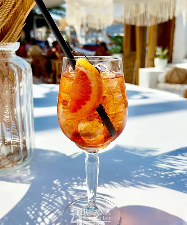Reana Beach & Bar Gölköy'ün beyaz masasında duran, üzerinde portakal dilimi, buz ve siyah pipet bulunan turuncu renkli ferahlatıcı kokteyl, arkada görünen yazlık plaj ve bar ambiyansıyla Bodrum'da dinlendirici bir atmosfer sunuyor.