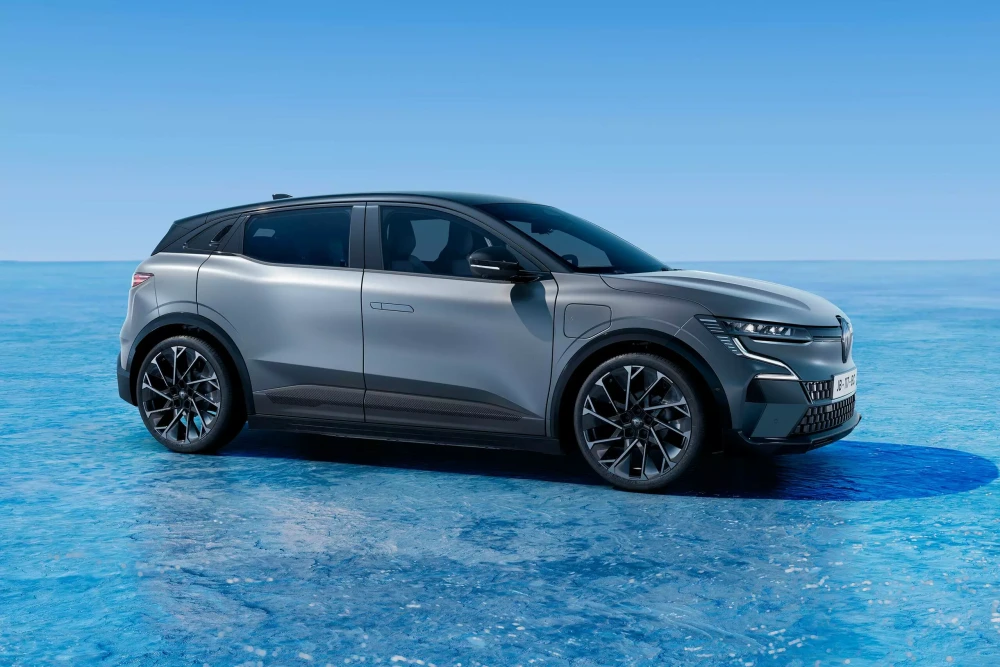 Gümüş-gri renkteki, modern ve sportif hatlara sahip Renault Megane E-Tech elektrikli compact otomobil, şarj portu görünür bir şekilde, açık mavi renkli, dalgalı bir yüzey üzerinde konumlanmış olup, aracın dinamik dış mekan tasarımını ve minimalist çizgilerini gözler önüne seriyor.