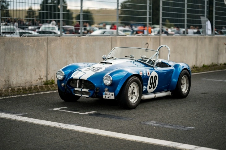 Mavi-beyaz çizgili Shelby Cobra 427, pistte yarış numarası 98 ile park halinde. Hafif gövdesi ve güçlü V8 motoruyla efsaneleşmiş klasik yarış otomobili.