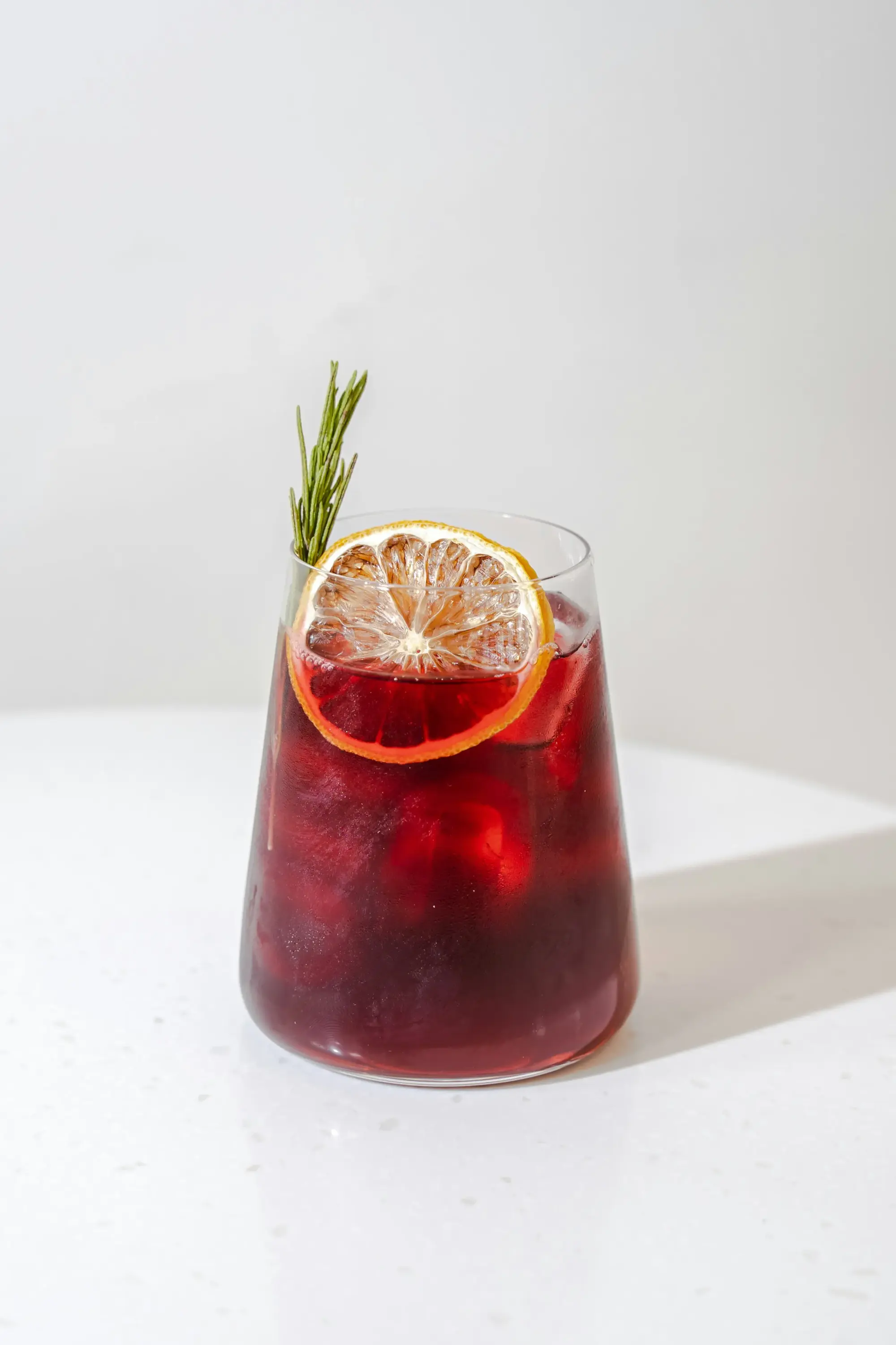 Buzlu hibiskus çayı, portakal dilimi ve biberiye ile süslenmiş kırmızı soğuk bitki çayı bardağı.