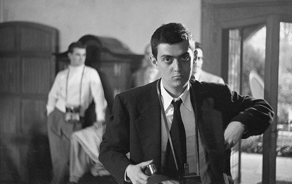 Genç Stanley Kubrick, koyu renk takım elbise, beyaz gömlek ve kravatıyla ciddi bir ifadeyle doğrudan kameraya bakarken, boynunda ve elinde kameralarıyla bir fotoğrafçı olarak ilk yıllarını yansıtan loş bir iç mekanda duruyor; arka planda ise bulanık diğer insanlar ve mobilyalar seçiliyor.