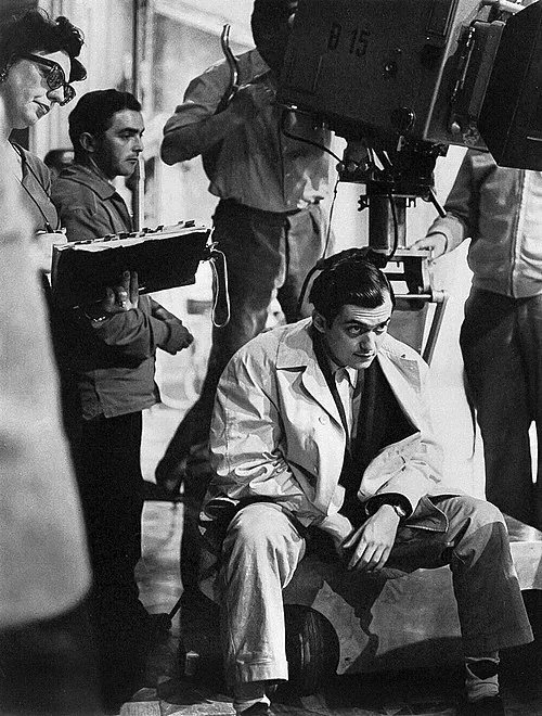 Siyah beyaz çekimde, bir film setinde, etrafında çalışan ekibi ve büyük bir kamera varken, düşünceli bir şekilde alçak bir tabureye oturmuş trençkotlu genç Stanley Kubrick, kendine özgü sinematik dehasını yansıtan bir sahne yönetiyor gibi görünüyor.
