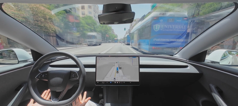 Otonom sürüş deneyimini vurgulayan Tesla Model Y elektrikli SUV otomobilin modern iç mekan tasarımı, ellerini direksiyona hafifçe koymuş sürücünün bakış açısından, merkezi ekranda görünen trafik arayüzü ve dışarıdaki şehir caddesindeki otobüsler ile kamyonu sergiliyor.
