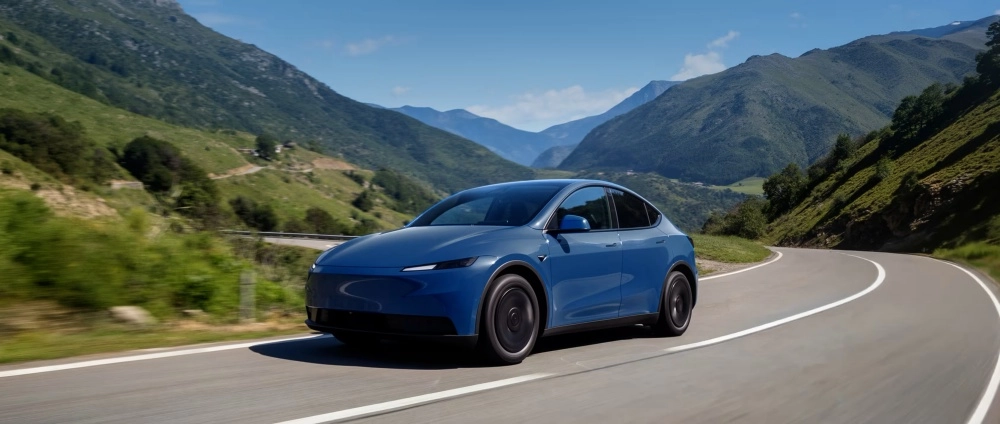 Mavi renkteki bir Tesla Model Y standart arkadan itişli elektrikli SUV, güneşli bir günde yeşil dağlık arazide virajlı bir yolda dinamik bir şekilde ilerlerken, zarif dış mekan tasarımını gözler önüne seriyor.