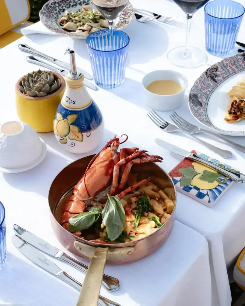 Bir trattoria masasında, beyaz örtü üzerinde duran bakır tencerede bol yeşillik ve fesleğenle servis edilmiş büyük bir ıstakoz ve gnocchi tabağı; etrafında mavi yivli bardaklar, desenli porselen tabaklar ve üzerinde Olio yazan limon desenli seramik zeytinyağı şişesiyle zengin bir İtalyan ziyafetini gösteren canlı bir görünüm.