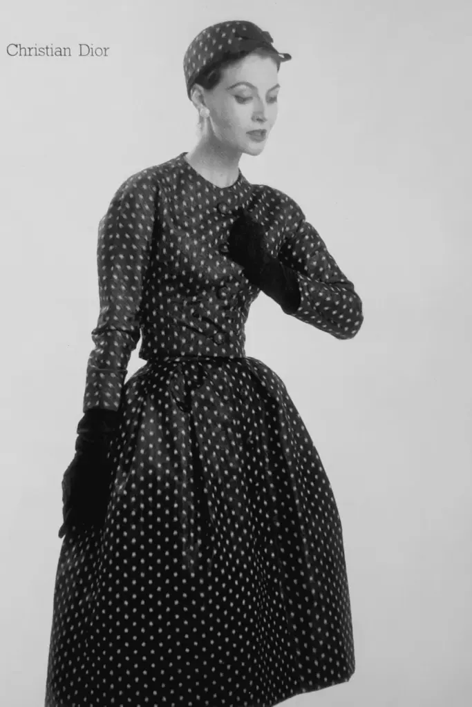 Görsel, Christian Dior markasına ait, 1950'lerin feminen ve zarif tarzını yansıtan, boydan boya küçük beyaz puantiye deseniyle kaplı, uzun kollu, vücuda oturan üst kısmı düğmeli ve kabarık etekli siyah bir elbise giymiş, eldivenli ve şapkalı bir kadın modeli siyah beyaz olarak sunuyor.