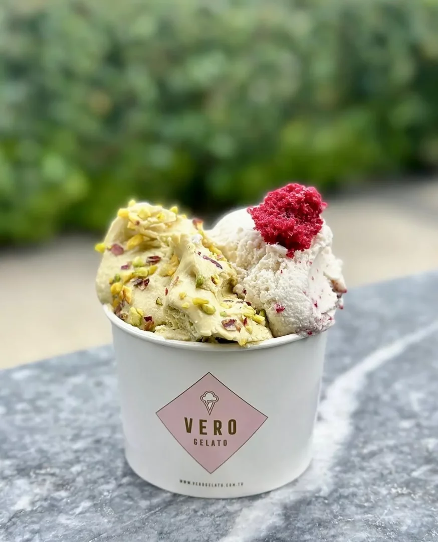 Vero Gelato logolu beyaz karton kapta, üzeri antep fıstığı parçacıklarıyla zenginleştirilmiş fıstıklı gelato ve kırmızı meyve parçacıklı beyaz gelato topları, en üstte parlak kırmızı bir crumble ile taçlandırılmış şekilde gri mermer bir yüzeyde, yeşil bitki örtüsü arka planında iştah açıcı ve ferahlatıcı bir görünüm sergiliyor.