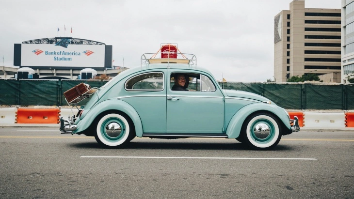 Açık mavi klasik Volkswagen Beetle, şehirde yol alırken yan profilden görüntüleniyor. Tavanında bagaj rafı ve valizlerle nostaljik bir görünüm sergiliyor.