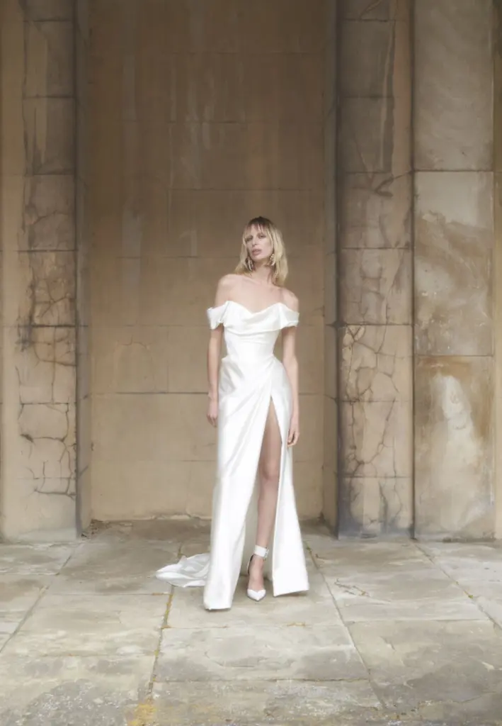 Off-shoulder drapeli saten kumaştan, bacak yırtmaçlı modern gelinlik. Minimal çizgileri ve zarif siluetiyle 2026 gelinlik trendlerini yansıtan çağdaş bridal tasarım.