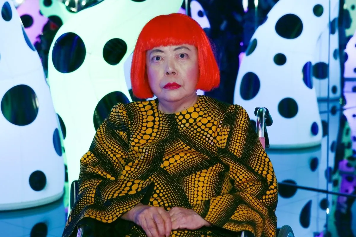 Kırmızı peruklu ve kendi imzası haline gelmiş, farklı boyutlardaki sarı-siyah puantiyeli desenlerle bezeli elbiseli avant-garde sanatçı Yayoi Kusama, arka planda ışıklı ve büyük siyah puantiyeli beyaz enstalasyon eserlerinin arasında tekerlekli sandalyesinde doğrudan bakıyor.