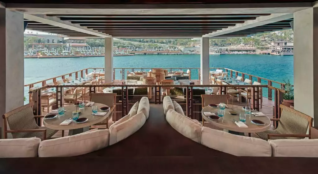 Ahşap masaların özenle düzenlendiği, denize nazır Bodrum'daki bu lüks mekanda, beyaz oturma grupları ve hasır sandalyeler eşliğinde, berrak turkuaz sulara karşı eşsiz bir yemek deneyimi vaat ediliyor.