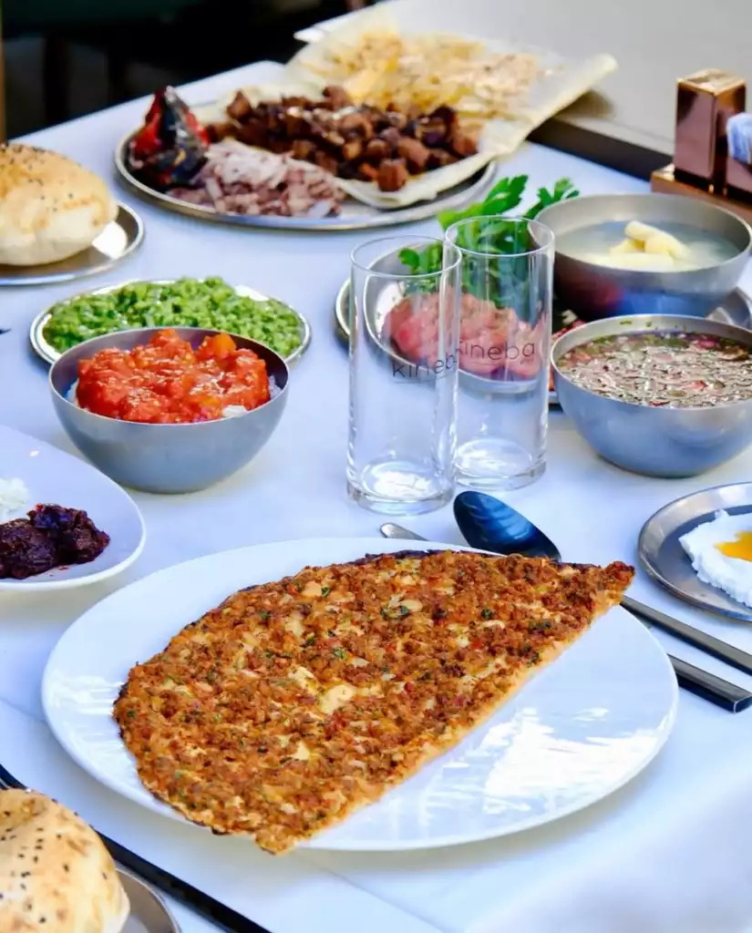 Ön planda taze pişmiş, bol kıymalı lahmacunun beyaz bir tabakta iştah açıcı duruşuyla, arka planda çeşitli mezeler, et yemekleri, çorbalar ve şişkin ekmeklerle dolu zengin bir Türk yemek sofrası gözler önüne seriliyor.