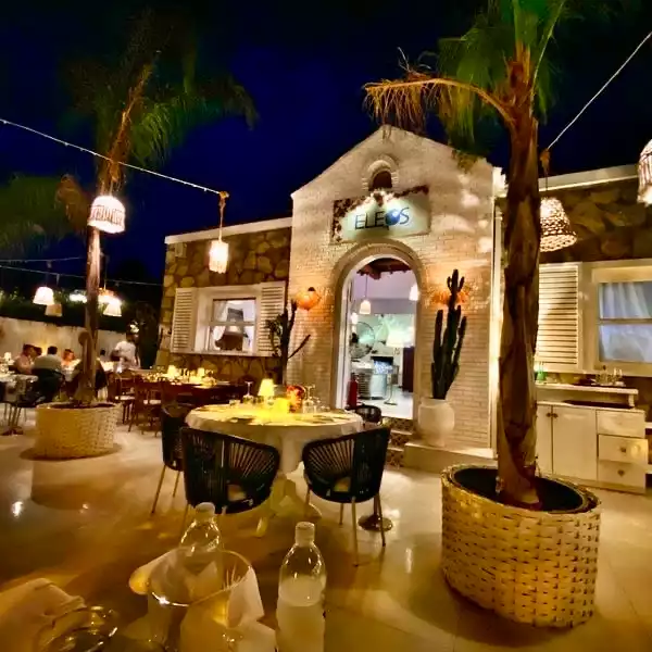 Eleos adlı şık restoranın palmiye ağaçları, romantik asma lambalar ve beyaz tuğla mimarisiyle aydınlatılmış, misafirlerin samimi bir atmosferde keyifle yemek yediği büyüleyici gece açık hava yemek alanı.
