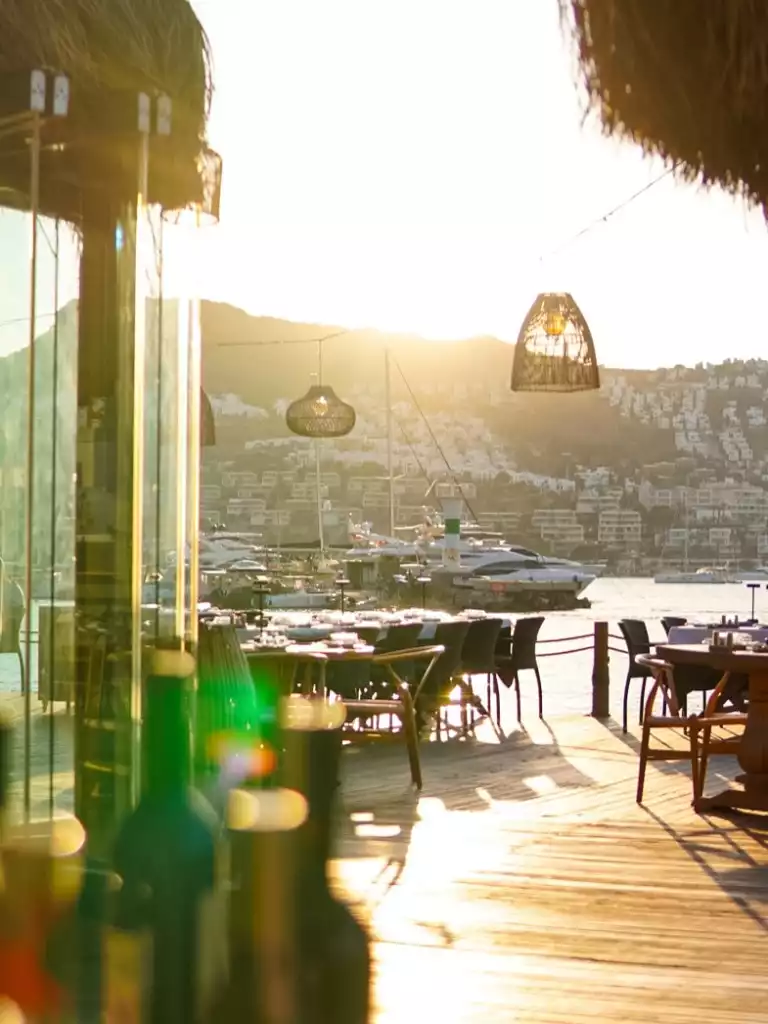 Bodrum'un eşsiz güzellikteki gün batımını seyreden, ahşap bir iskele üzerine kurulmuş lüks bir deniz kenarı restoranda, masalar ve hasır lambalar altın rengi ışıklarla aydınlanırken, arka planda yatlar ve tepelerdeki beyaz evler görünüyor.