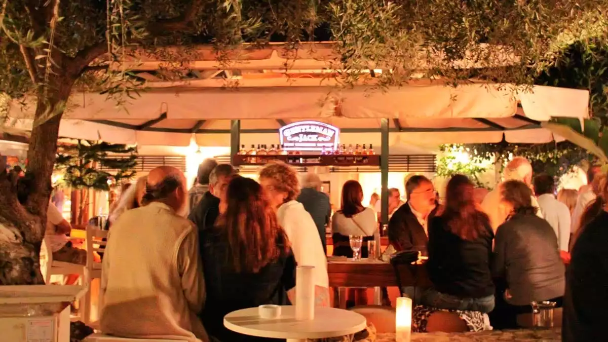 Bodrum'un hareketli restoranlarından birinde, zeytin ağaçlarının yeşil yaprakları altındaki sıcak bir akşamda, aydınlatılmış bir dış mekan barda sosyalleşen ve keyifli anlar yaşayan kalabalık bir insan topluluğu görülüyor.