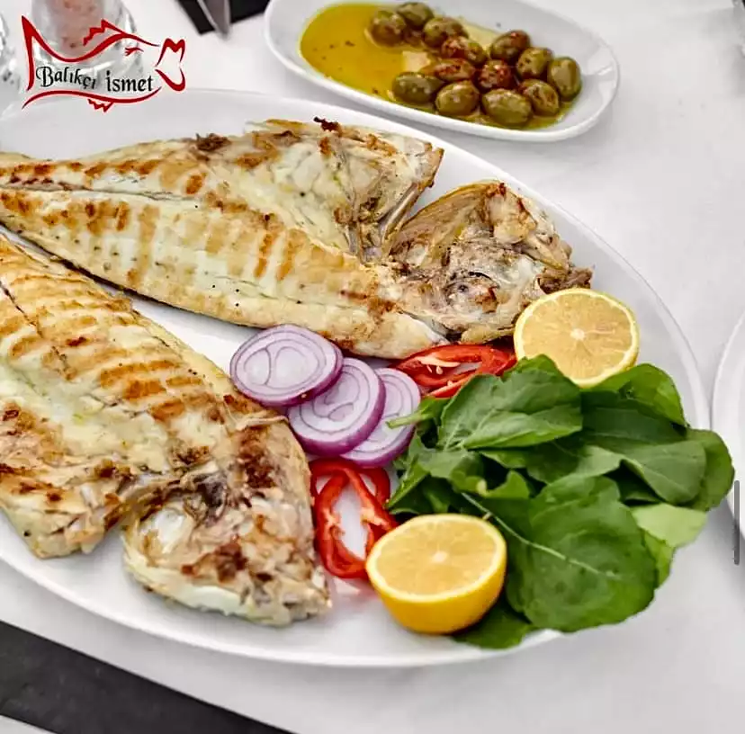 Bodrum'da, Balıkçı İsmet restoranında, ızgara balık keyfini yansıtan bu karede, taze yeşillikler, mor soğan halkaları, kırmızı biber dilimleri ve dilimlenmiş limonla sunulan iki bütün ızgara balık, beyaz oval bir tabağı dolduruyor; arka planda zeytin ve zeytinyağı içeren küçük bir kase belirginleşiyor.